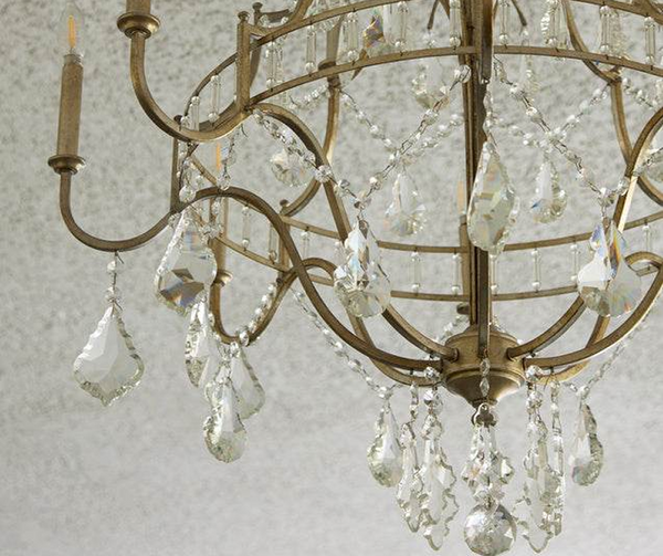 A gold-framed chandelier with curved arms and hanging crystal drops, photographed against a soft, light background