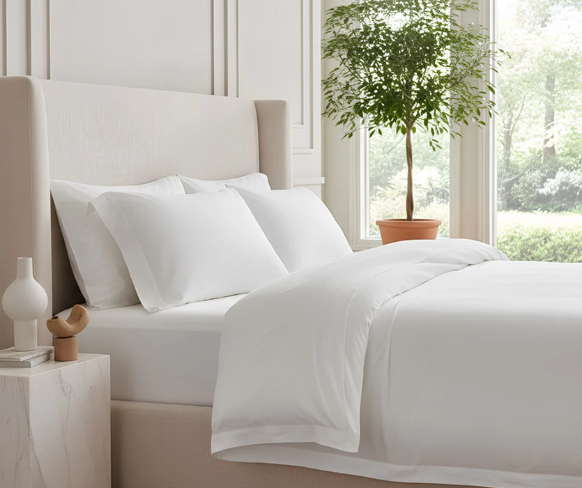 Bed dressed in crisp white linens with a cream upholstered headboard and a bedside table in a bright, softly lit, neutral bedroom