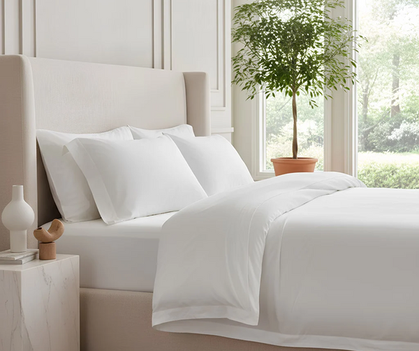 Bed dressed in crisp white linens with a cream upholstered headboard and a bedside table in a bright, softly lit, neutral bedroom