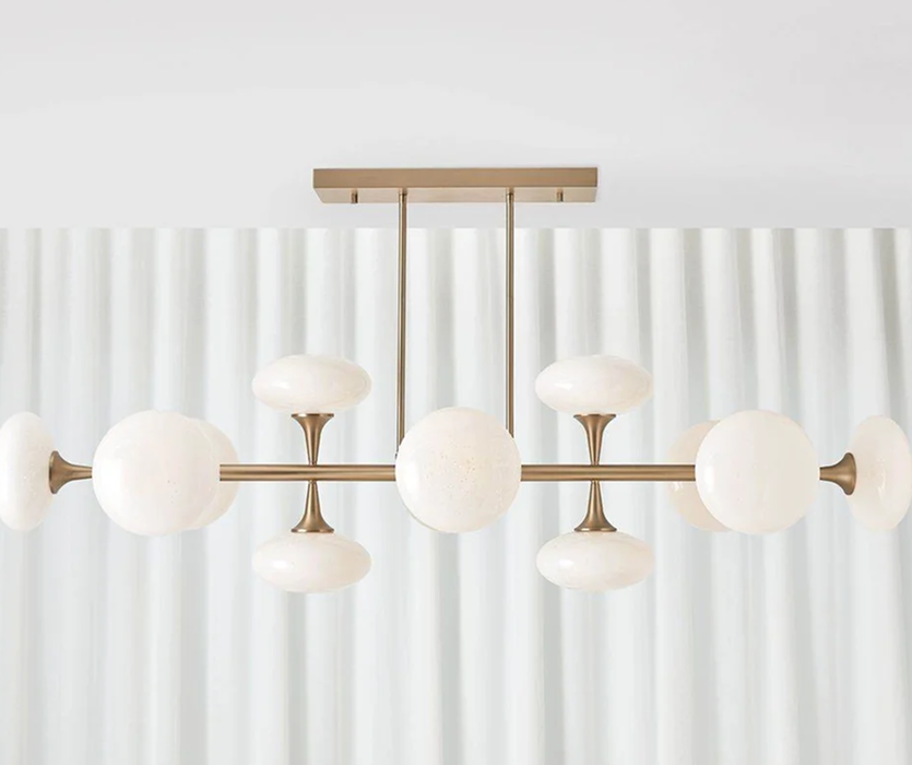 Modern brass chandelier with branching arms and round white glass globes, hanging in front of sheer white curtains in a bright room