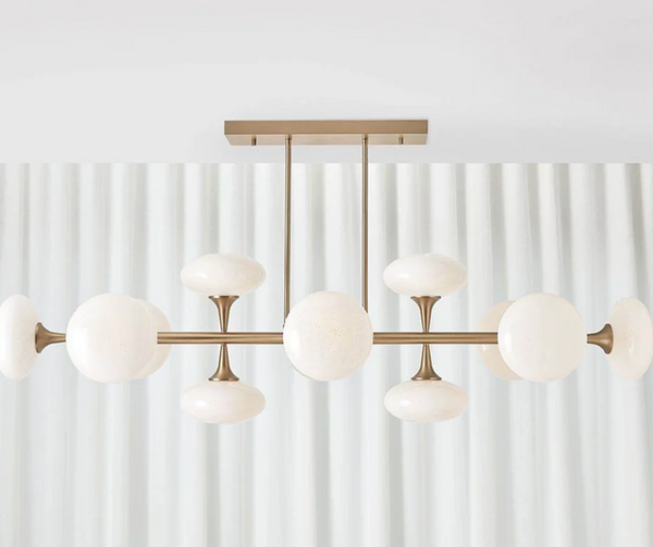 Modern brass chandelier with branching arms and round white glass globes, hanging in front of sheer white curtains in a bright room