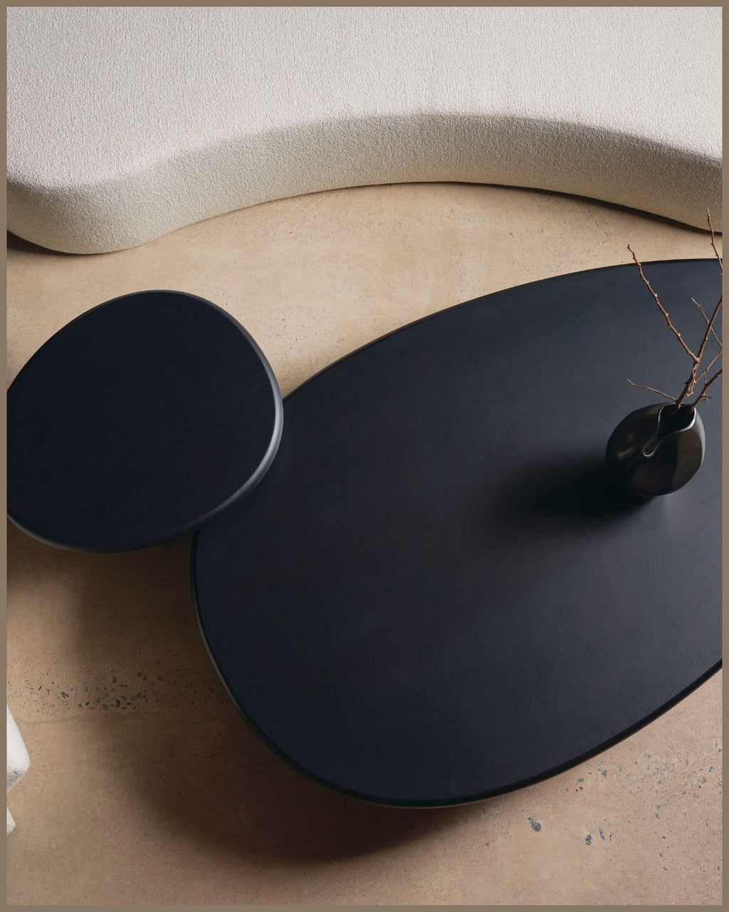 A modern black coffee table with an organic organic-shaped main surface and a round matching side table, styled in a minimalist interior with a textured beige floor and a cream curved sofa.