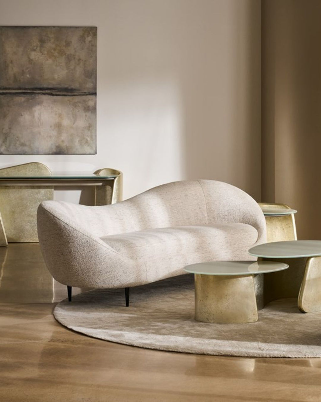 A modern curved upholstered sofa in a warm beige fabric, styled in a minimalist living room with matching round side tables, a textured rug, and a neutral-toned wall.