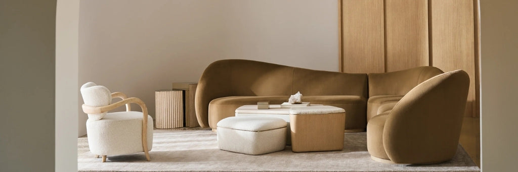 A modern living room furniture set featuring a curved brown velvet sofa, a matching armchair, a white accent chair, and a low rectangular coffee table with a white ottoman, set against a beige wall and light wood paneling.