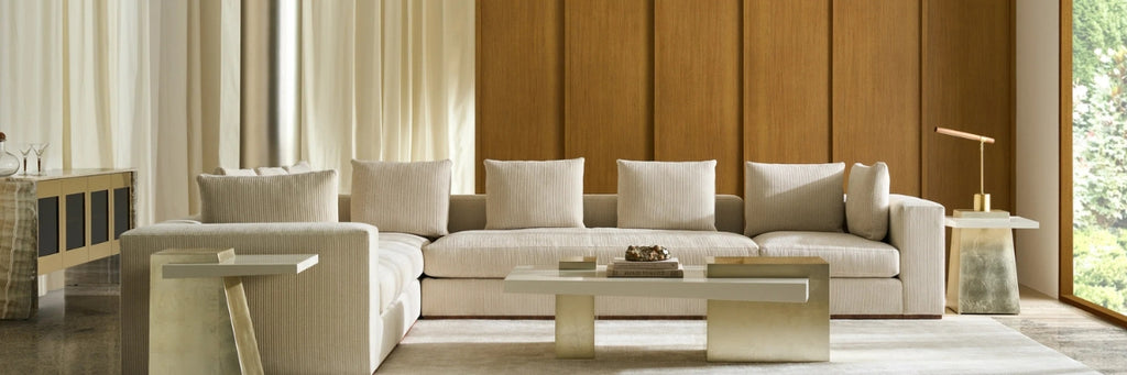A modern sectional sofa in a warm cream or off-white fabric, styled in a contemporary living room with a wooden paneled wall, a low rectangular coffee table, and side tables.
