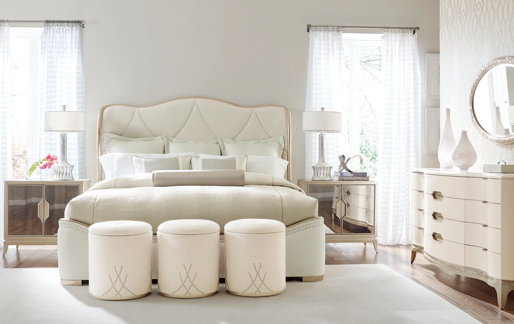 Lit capitonné incurvé en ivoire avec tête de lit à panneau arqué et trois poufs ronds cloutés à côté de tables de chevet miroir dans une chambre de luxe entièrement blanche