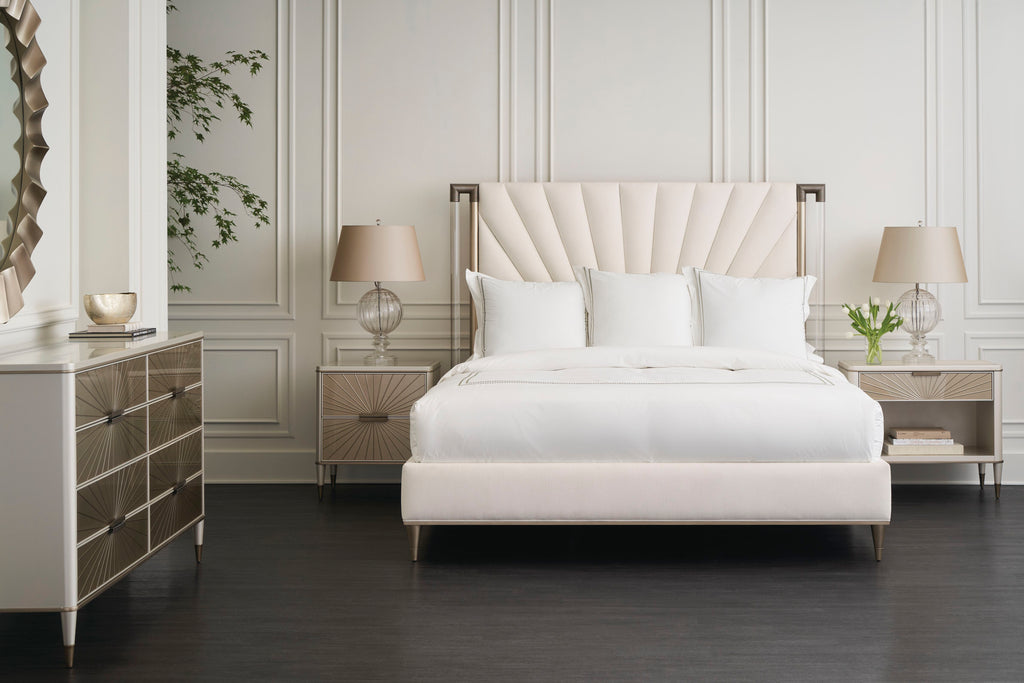 Lit tapissé à plis éventail crème avec embouts de poteau en bronze, à côté de tables de chevet à tiroirs "rayons de soleil" et d'une commode assortie dans une chambre à panneaux blancs.