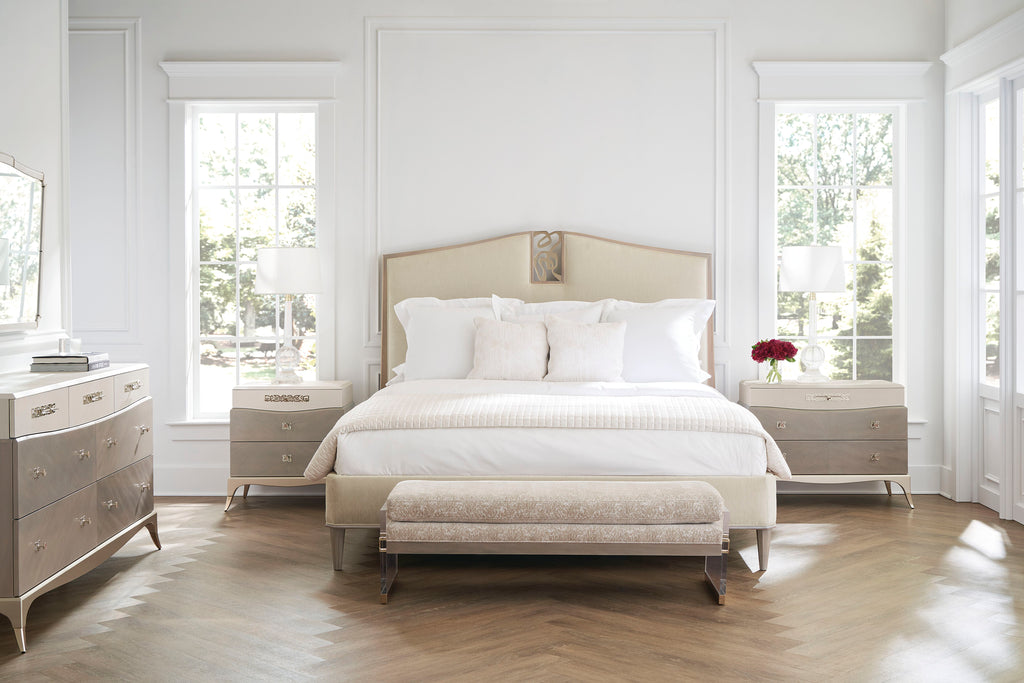 Lit rembourré beige champagne avec tête de lit arquée en dos d'âne et tables de chevet incurvées à trois tiroirs assorties et banc sur un parquet à chevrons.